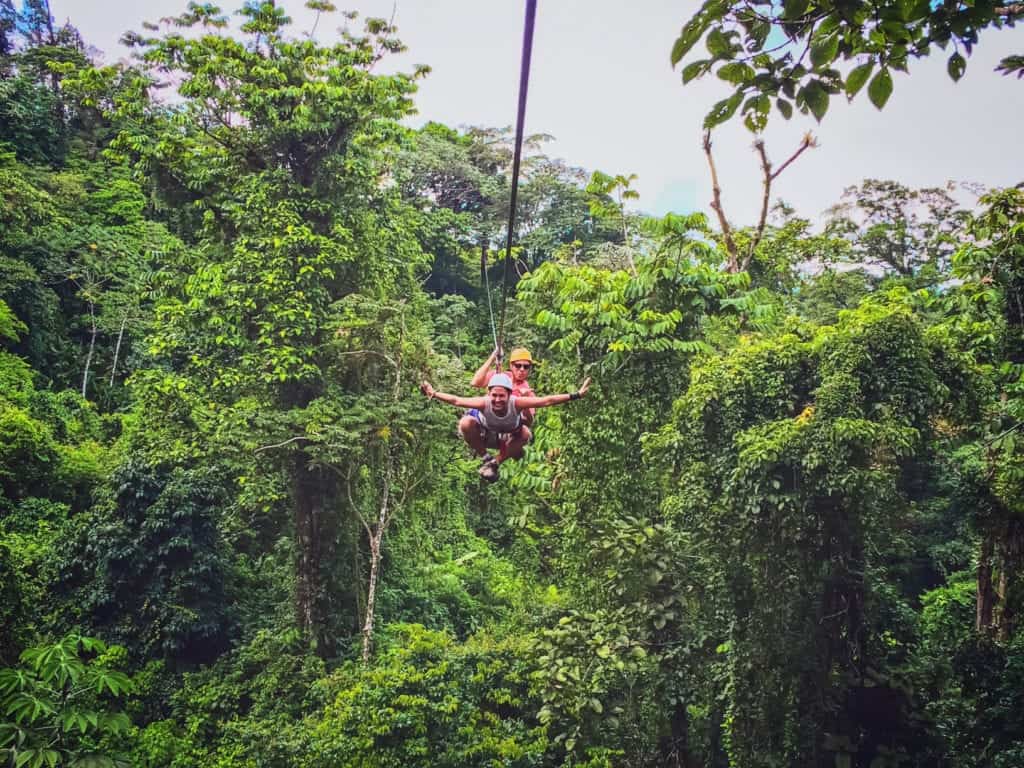 People riding a zipline over green rain forest while smiling towards the cameraman.